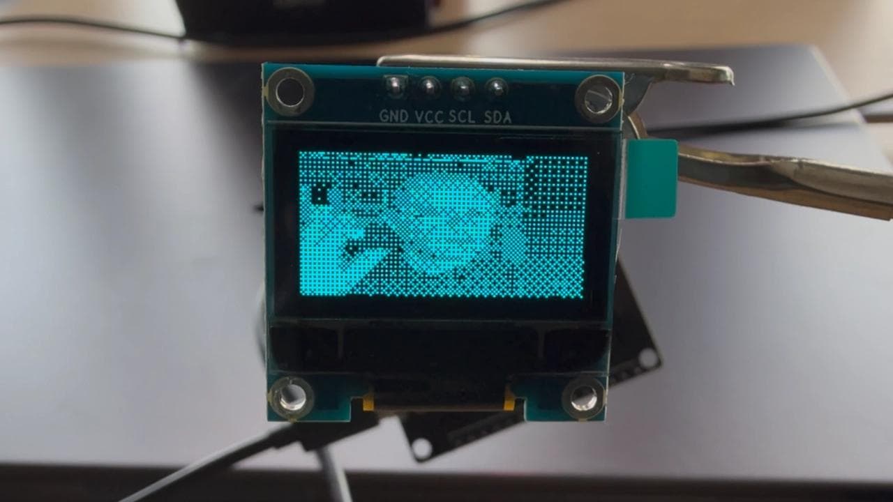 A tiny OLED display connected to an ESP32 showing a live webcam feed in a dithered 1-bit pixel style, streamed wirelessly from the browser over Wi-Fi.
